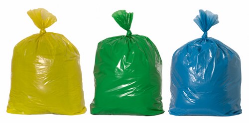 The image shows three large plastic rubbish bags placed on a paved outdoor surface, likely part of a waste collection or disposal process. The bags are made of glossy, durable plastic material and are filled with waste, giving them a rounded, bulky appearance. The bag on the left is bright yellow, positioned upright with a twisted top sealed with a knot. The central bag is green, also tied at the top, and stands slightly to the right of the yellow bag. The third bag, on the far right, is blue and similarly secured at the top, with a slightly crumpled texture. The background appears to be a paved area in an urban or industrial environment, consistent with a location in Whitechapel or nearby areas where businesses might handle waste collection. The lighting is natural, indicating daytime, with no visible vehicles or equipment in the scene. This image exemplifies typical waste packaging used in commercial rubbish removal services provided by Business Waste Removal Whitechapel, highlighting the preparation for waste pickup or disposal in the local borough.