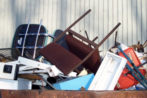 A pile of discarded household and office waste items stacked outdoors against a corrugated metal wall, including a dark brown fabric-upholstered armchair with wooden legs lying on its side, a white plastic storage container, a blue plastic bin, a red plastic chair, and various metal and plastic furniture parts. The items appear weathered and are arranged in a haphazard manner, suggesting they are awaiting rubbish removal. The environment resembles a residential or commercial area with concrete pavement visible at the base of the pile, indicating a typical urban setting in Whitechapel, East London. This scene illustrates the type of clutter that Business Waste Removal Whitechapel services in accordance with local waste management regulations, focusing on clearing bulky or unwanted household and business rubbish from the area.