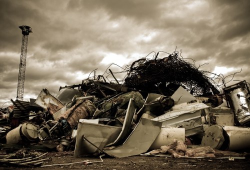 A large pile of discarded household and industrial debris situated outdoors on a dirt surface, consisting of broken appliances such as washing machines and refrigerators with visible metal and plastic panels in worn, chipped, and faded finishes. The debris also includes twisted metal parts, scattered wires, and unidentifiable scraps, all piled haphazardly with some items leaning against each other. In the background, a metal telecommunications tower stands to the left under a cloudy, overcast sky, casting a muted lighting over the scene. The environment suggests an industrial or waste management site, likely in an area near Whitechapel, where Business Waste Removal Whitechapel offers rubbish clearance services. The mixture of textures from smooth metal surfaces and rough, weathered plastics highlights the complex composition of waste materials typically handled in rubbish removal operations.