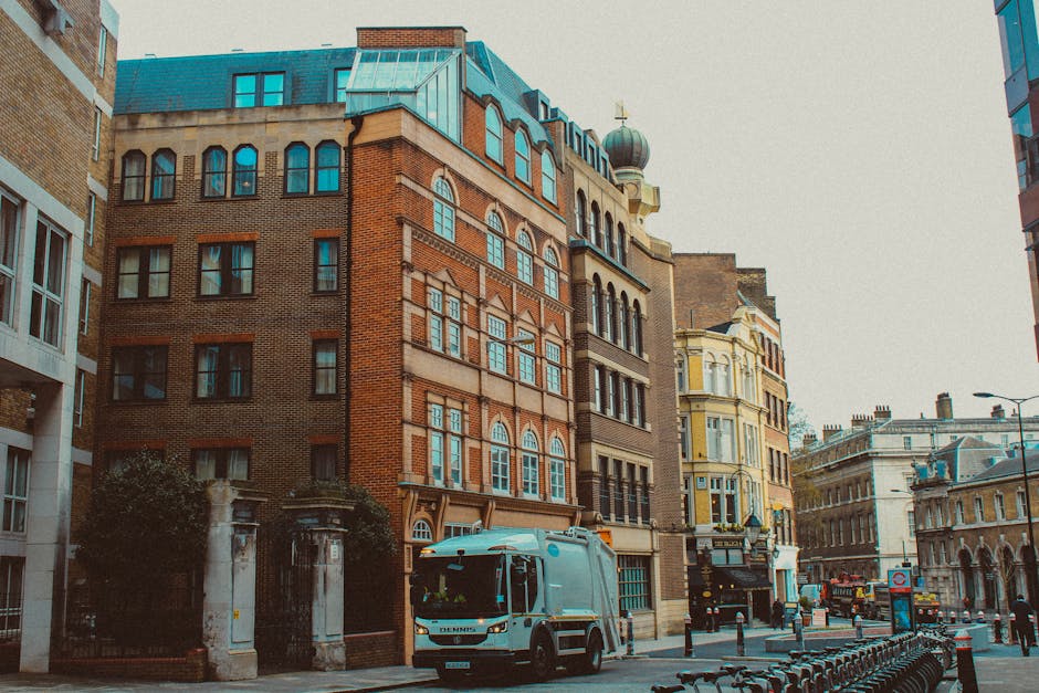 A city street scene featuring a row of multi-storey buildings with diverse architectural styles and facades, including red brick, stone, and ornate detailing. The buildings have large, arched windows 